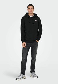 Zwarte hoodie met koordjes en een voorkraagzak, gecombineerd met donkere jeans en grijze sneakers, staand tegen een lichte achtergrond.