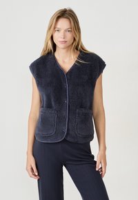 Kobieta w granatowej futrzanej kamizelce bez rękawów z kieszeniami z przodu i guzikami, w połączeniu z ciemnymi prążkowanymi spodniami na jednolitym tle.