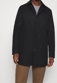 Svart knäppjacka med krage, med rak skärning och sidofickor. Bärs över en randig skjorta. Slät tygstruktur.