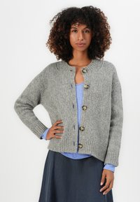 Woman with curly hair wearing a gray chunky knit buttoned cardigan over a blue top and dark skirt, standing with one hand on hip.