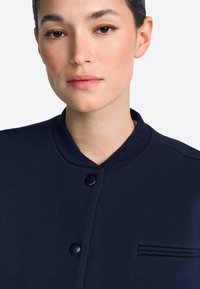 Marineblauw bomberjack met geribde kraag, sluiting met twee knopen en een decoratieve zakaccent. Soepele textuur en een getailleerd ontwerp.