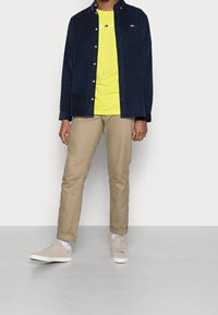 Tmavě modrá manšestrová košile s knoflíky přes jasně žluté tričko, béžové chinos kalhoty a světle zbarvené tenisky s viditelným prošitím.