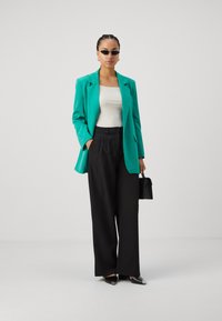 Ein türkisfarbener Blazer über einem weißen Oberteil, kombiniert mit schwarzen weit geschnittenen Hosen. Als Accessoires dienen kleine schwarze Sonnenbrillen und eine strukturierte Handtasche.