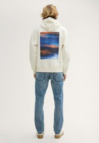 Sudadera crema con estampado gráfico en azul y naranja en la parte trasera. Combinada con jeans azul claro y zapatillas beige. Ajuste sencillo y relajado.