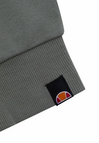 Puño de sudadera de punto gris que presenta una etiqueta negra con un logotipo de color, mostrando un semicírculo con acentos en naranja, rojo y blanco.