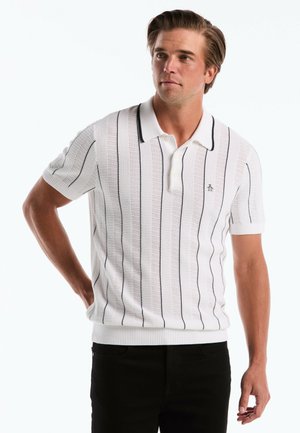 Junger Mann trägt ein weißes, kurzärmeliges Poloshirt aus Strick mit dünnen schwarzen vertikalen Streifen, steht mit einer Hand in der Hüfte.