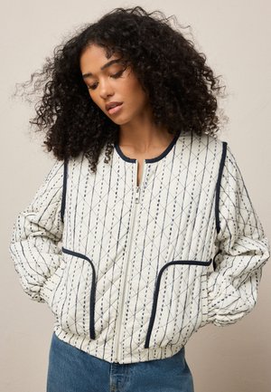 Femme aux cheveux bouclés portant une veste matelassée blanche zippée avec des lignes bleues surpiquées et un biais bleu marine, les mains dans les poches, regardant vers le bas.