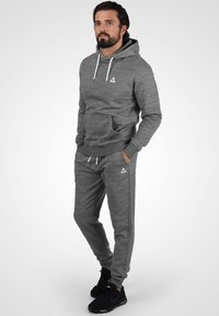 Sudadera gris con cordones blancos en contraste y logotipo. Combinada con pantalones de chándal grises con franjas laterales y bolsillos. Zapatillas negras.