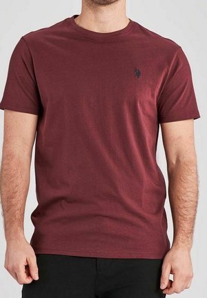 Mand iført simpel mørkerød kortærmet t-shirt med lille mørkt logo på venstre bryst, kombineret med sorte bukser, mod ensfarvet baggrund.