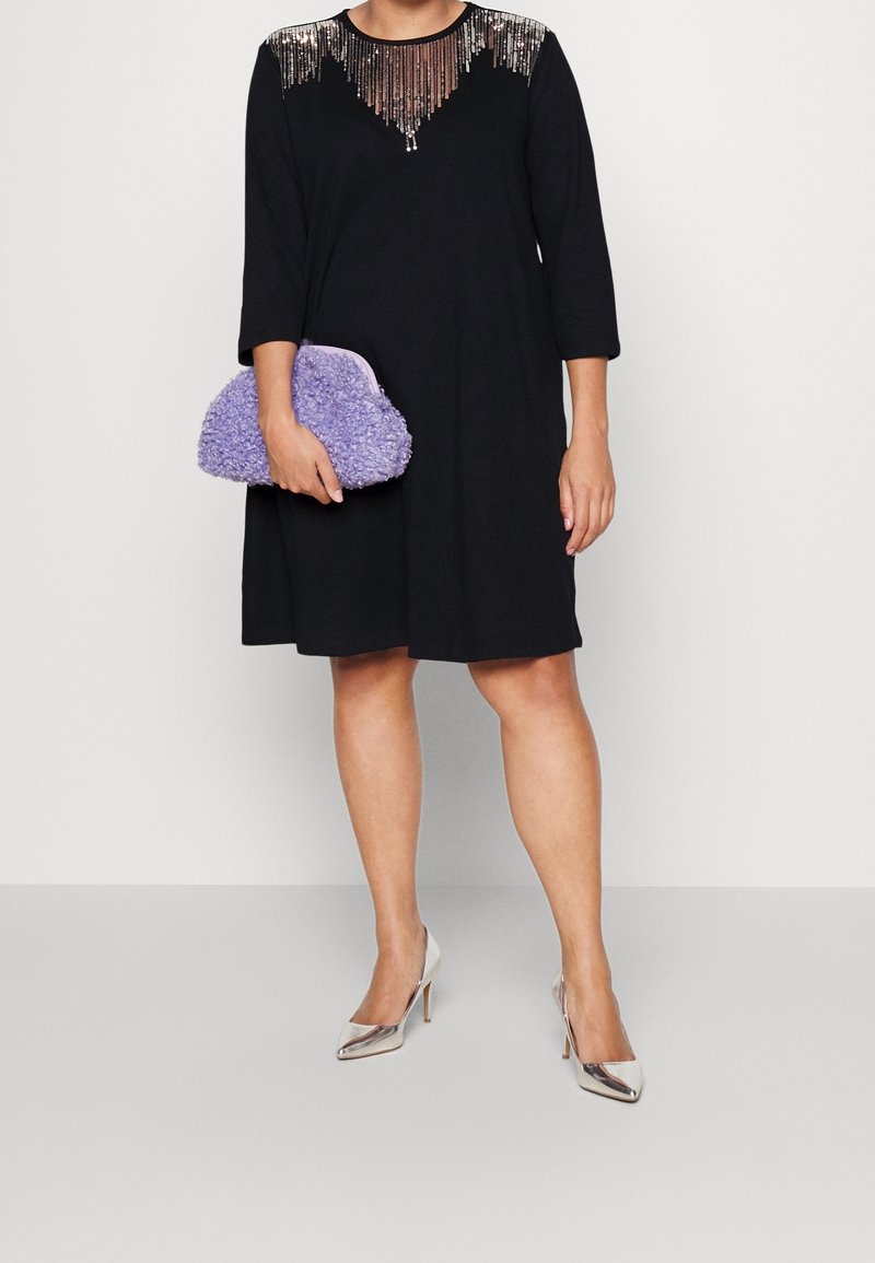 Svart klänning med en paljettprydd halsringning, trekvartslånga ärmar. Matchad med en texturerad lila kuvertväska och silvriga högklackade skor.