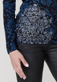 Top de manga larga con un patrón floral azul, adornado con bordados plateados y lentejuelas, combinado con pantalones de cuero negros.
