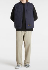 Veste bleu marine avec col, fermeture éclair à l'avant et deux poches poitrine ; portée sur une chemise noire, associée à un pantalon beige et des chaussures slip-on à carreaux.