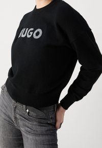 Schwarzer Pullover mit einem großen, silbernen "HUGO"-Logo auf der Vorderseite, klassischen Rundhalsausschnitt und lässigen Ärmeln. Kombiniert mit dunkelgrauen Jeans.