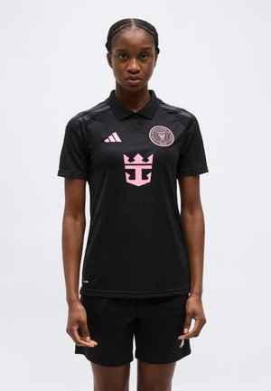 Ženska stoji obrnjena naprej, oblečena v črn nogometni dres Miami Inter Miami CF z roza logotipi in črne Adidas kratke hlače.