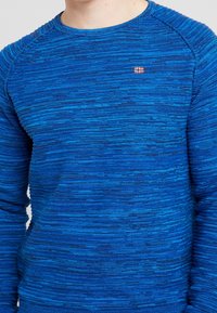 Blaues strukturiertes Strick-Sweatshirt mit Rundhalsausschnitt, das dezente horizontale Streifen und ein kleines rot-weißes Flaggenemblem auf der Brust trägt.