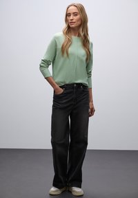 Hellgrüner Pullover mit langen Ärmeln, kombiniert mit schwarzer, weit geschnittener Jeans und beigen Sneakers, aus strukturiertem Material und mit lockerer Passform.