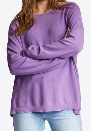 Femme portant un pull lavande à manches longues, les bras croisés, associé à un jean bleu clair, sur un fond blanc.