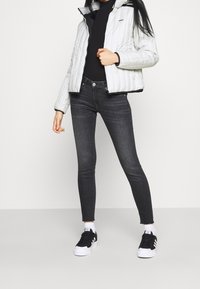 Mujer vestida con una chaqueta acolchada blanca, un suéter negro de cuello alto, jeans ajustados oscuros, calcetines blancos y zapatillas de deporte en blanco y negro, de pie contra un fondo liso.