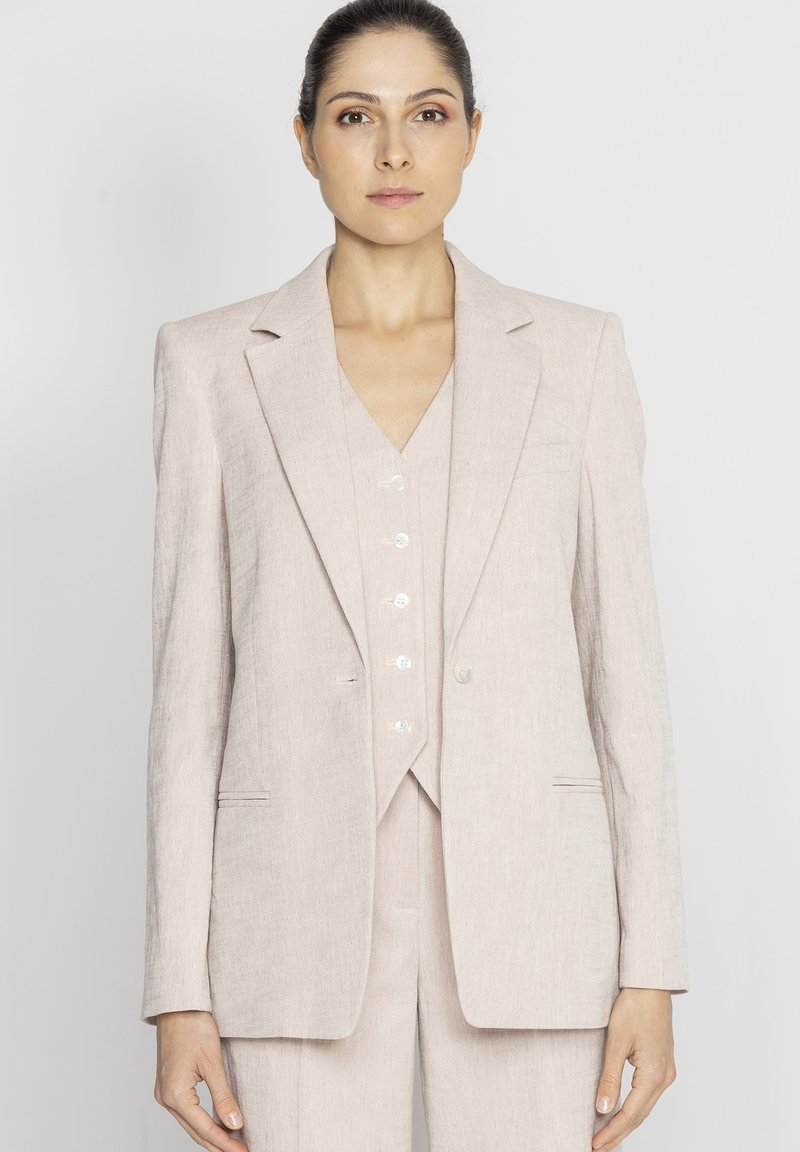 Jasnoróżowy garnitur z lnu, składający się z marynarki zapinanej na jeden guzik, z klapami i dopasowaną kamizelką z ozdobnymi guzikami. Gładka tekstura, dopasowany krój.