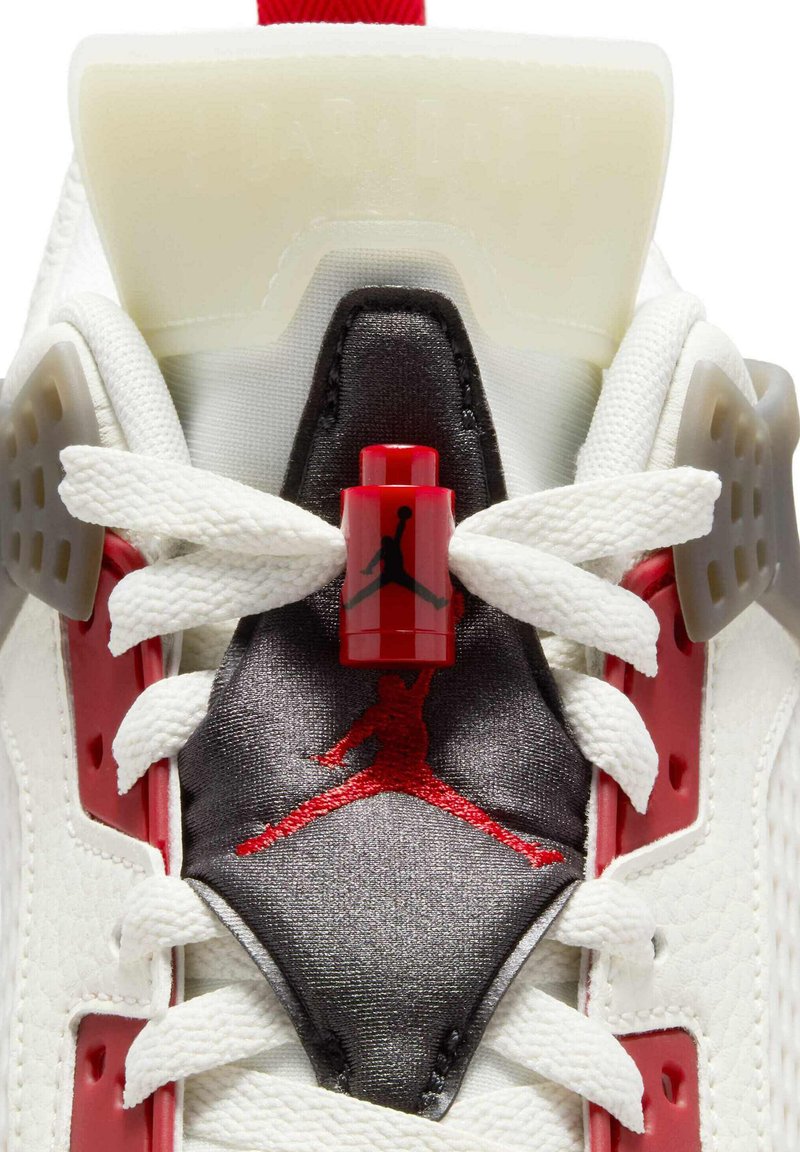 Zapatilla de baloncesto blanca con una lengüeta negra y roja con el logo de Jumpman, acentos rojos y superposiciones texturizadas. El sistema de bloqueo de cordones es rojo.