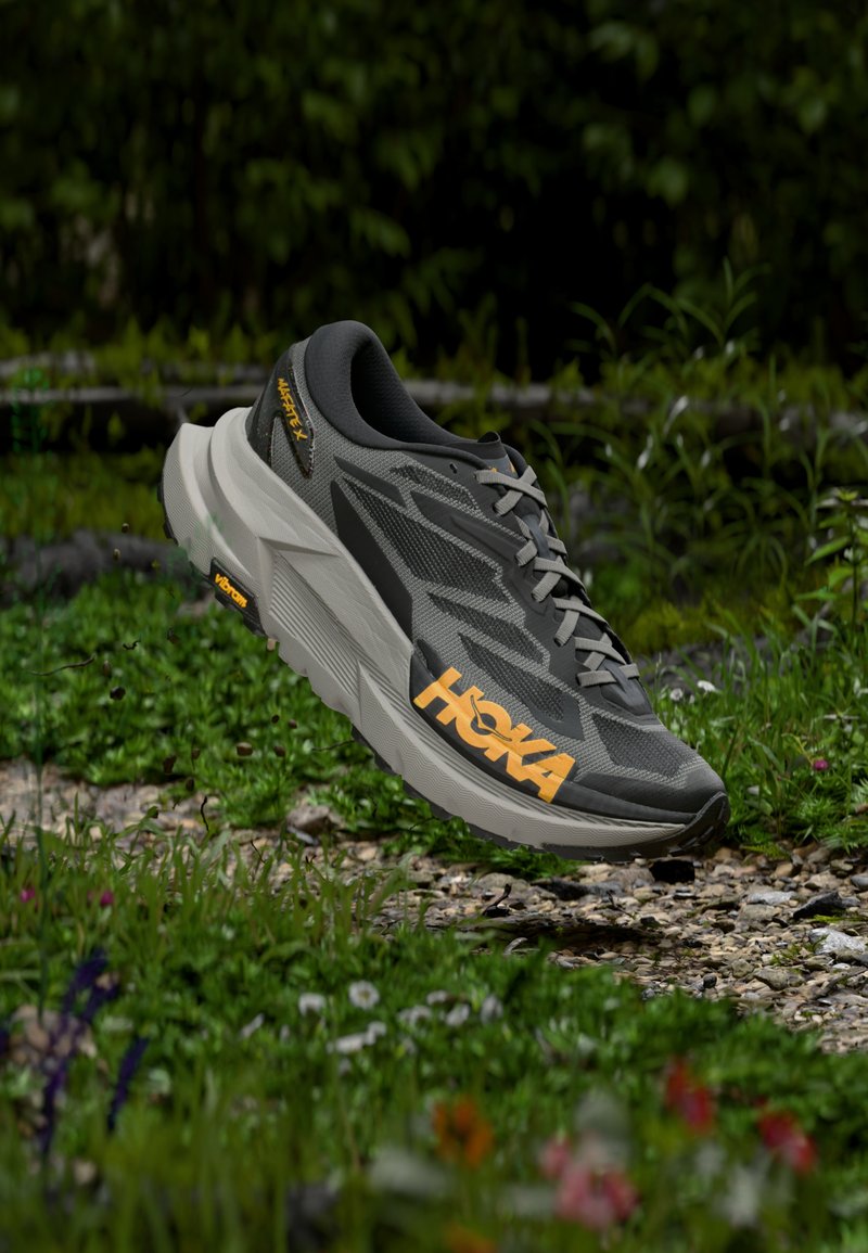 Γκρι και μαύρο παπούτσι τρεξίματος Hoka με πορτοκαλί λογότυπο να αιωρείται πάνω από έναν βραχώδη, πράσινο δασώδη δάπεδο με φυτά και λουλούδια.