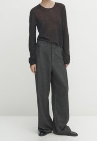 Person wearing a sheer black long-sleeve top, dark gray pinstripe wide-leg trousers, and black shoes, standing against a plain background.