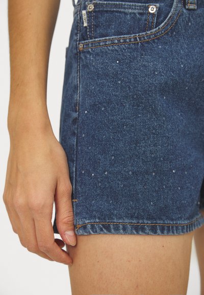 Shorts de mezclilla en color azul oscuro, diseño de cinco bolsillos y dobladillo deshilachado con pequeños adornos cerca de la superficie de la tela.