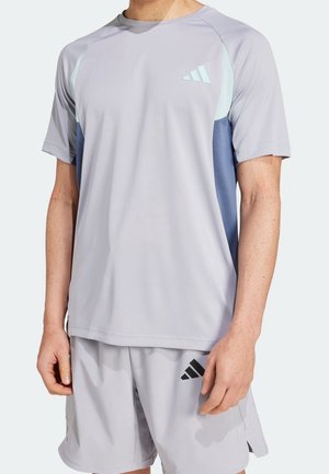 Homme portant un t-shirt de sport gris clair et un short assorti avec des logos Adidas gris foncé et bleu clair, visible du cou jusqu'à mi-cuisse.