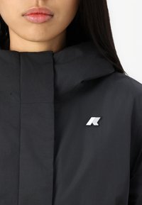 Černá kapuce bunda vyrobená z hladkého materiálu, se slušným bílým výšivkovým logem na prsou a zapínáním na zip. Jednoduchý a funkční design.