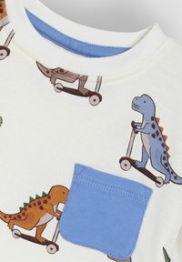 Valkoinen T-paita, jossa on värikkäitä dinosauruskuvastoja skoottereilla. Pyöreä kaula-aukko ja sininen tasku vasemmassa rintapielessä. Puuvillamateriaali.