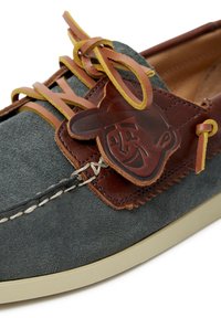 Zapato de barco de ante gris con acentos de cuero marrón, parche logo decorativo, suela beige y cordones amarillos. Detalles de costura a lo largo de los lados.
