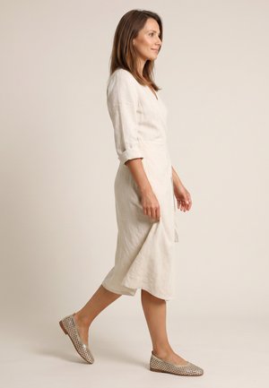 Kobieta idąca bokiem, ubrana w długą kremową sukienkę z podwiniętymi rękawami i srebrne, perforowane, płaskie buty na jednolitym beżowym tle.