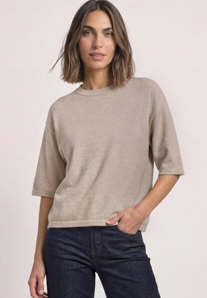 Femme aux cheveux mi-longs portant un haut en maille beige à manches courtes et un jean bleu foncé, main dans la poche, debout devant un fond uni.