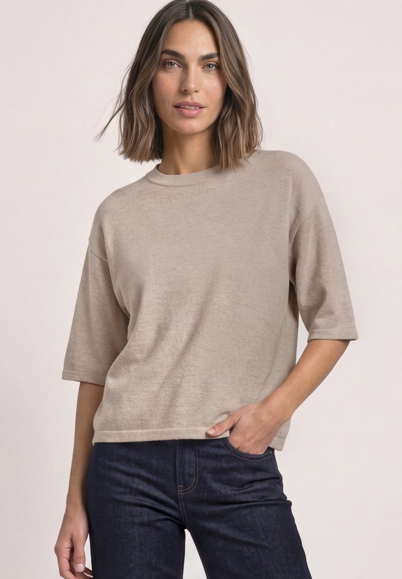 Femme aux cheveux mi-longs portant un haut en maille beige à manches courtes et un jean bleu foncé, main dans la poche, debout devant un fond uni.