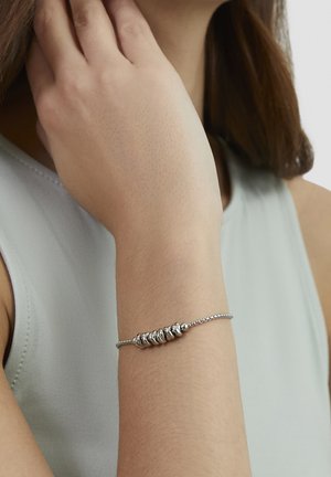 Frau trägt ein zartes silbernes Armband mit gruppierten, runden Perlen am Handgelenk, bekleidet mit einem ärmellosen, hellgrauen Oberteil.