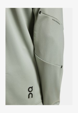 Světlé zelené sportovní sako s texturovaným povrchem, s kapsou na zip na rukávu a černým logem u lemu.