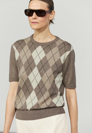 Kvinde iført store solbriller med skildpaddemønster og en brun og beige argyle kortærmet sweater med lyse bukser.