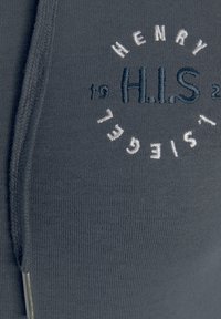 Tejido de sudadera gris oscuro con textura acanalada, con el logotipo bordado "H.I.S" y números en blanco y azul marino. Detalle de cremallera plateada.