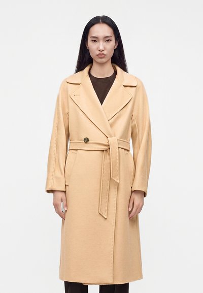 Femme aux longs cheveux noirs portant un manteau en laine beige ceinturé sur un haut foncé, debout devant un fond clair uni.