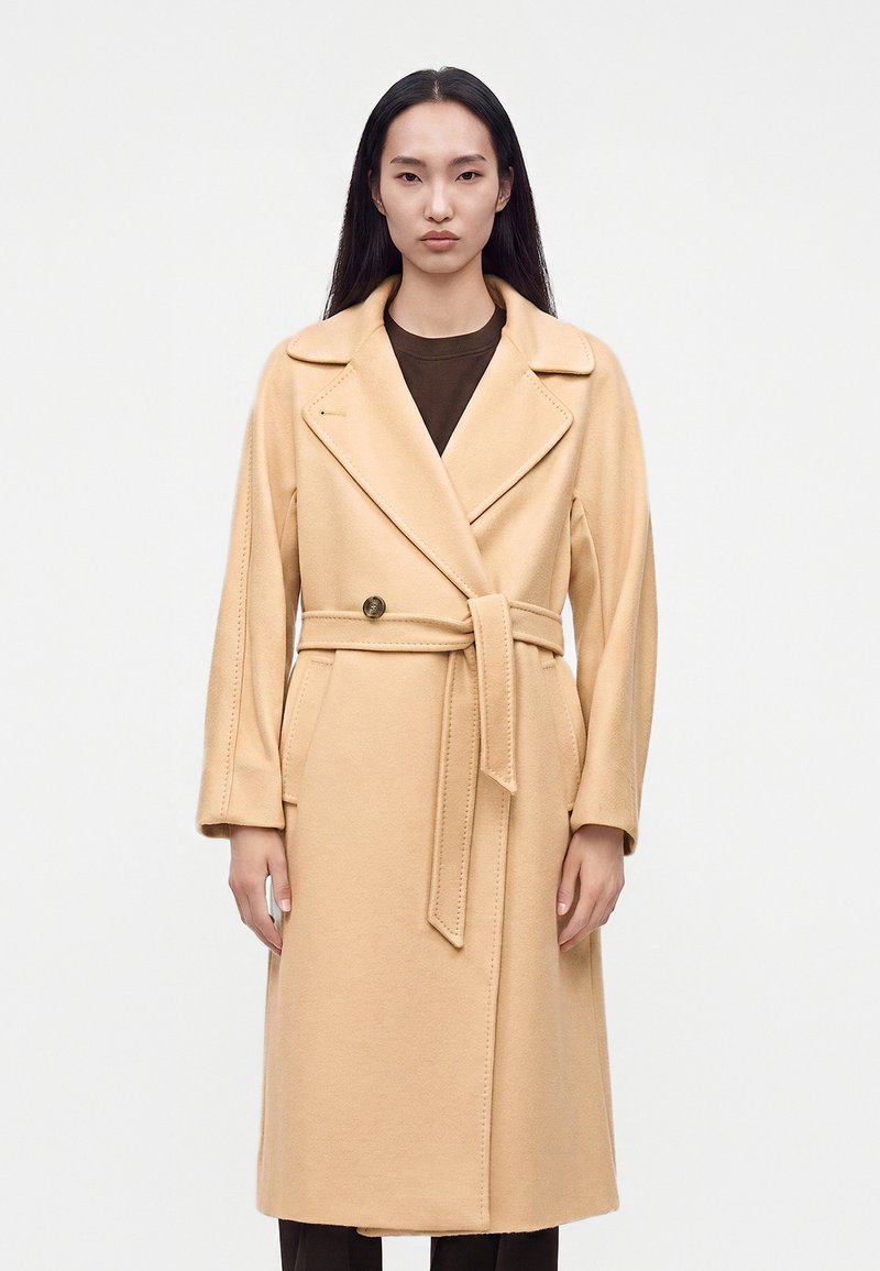 Femme aux longs cheveux noirs portant un manteau en laine beige ceinturé sur un haut foncé, debout devant un fond clair uni.