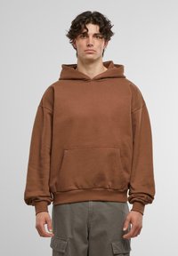 Hnědá mikina vyrobená z měkké tkaniny, s přední kapsou, volným střihem a kapucí na šňůrku. Jednoduchý design bez viditelných vzorů.
