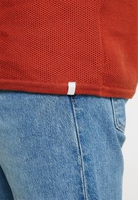 Red mesh short-sleeve shirt featuring a textured finish, with a visible white label at the hem, paired with light blue denim jeans.