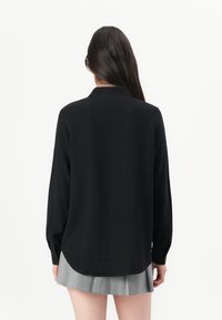 Zwarte blouse met lange mouwen en knoopmanchetten, voorzien van een ronde kraag en een gebogen zoom, gecombineerd met een grijze geplooide rok.