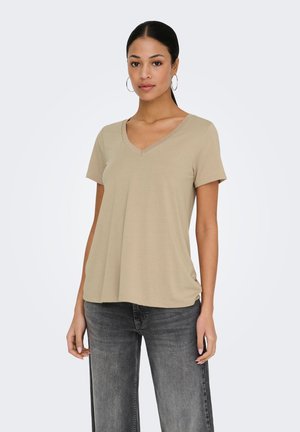 Femme portant des boucles d'oreilles créoles, un t-shirt beige à col en V à manches courtes et un jean noir délavé, debout devant un fond clair uni.