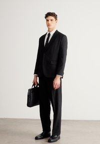 Czarny garnitur na miarę z marynarką i spodniami. Biała koszula i czarny krawat. Model trzyma czarną skórzaną teczkę, ma na sobie czarne buty.