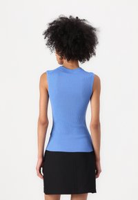 Woman seen from the back wearing a sleeveless ribbed blue top and a black skirt against a plain white background.