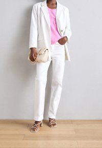 Blazer blanco sobre una blusa rosa, combinado con pantalones blancos y tacones de tiras. Un bolso crema con acentos de cadena completa el atuendo.