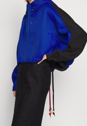 Person iført en blå tekstureret jakke med sorte ærmer og en sort nederdel med blomstret blondevæv, stående med den ene hånd på hoften.