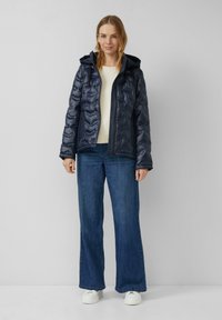 Femme debout portant une veste à capuche matelassée bleu marine, un pull crème, un jean large bleu et des baskets blanches à plateforme sur un fond uni.