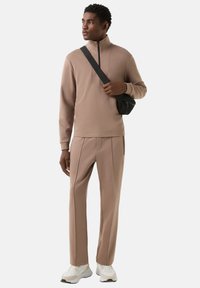 Světlě hnědá fleecová mikina na zip spárovaná se stejnobarevnými zužujícími se kalhotami s bočními švy, doplněná bílými sportovními botami a černou kabelkou přes rameno.